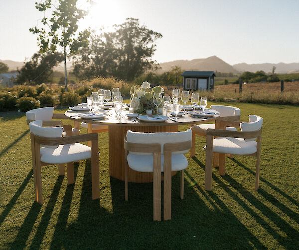 An outdoor dining setup with a round table, chairs, and tableware on a grassy lawn under a clear sky, surrounded by trees and distant hills.