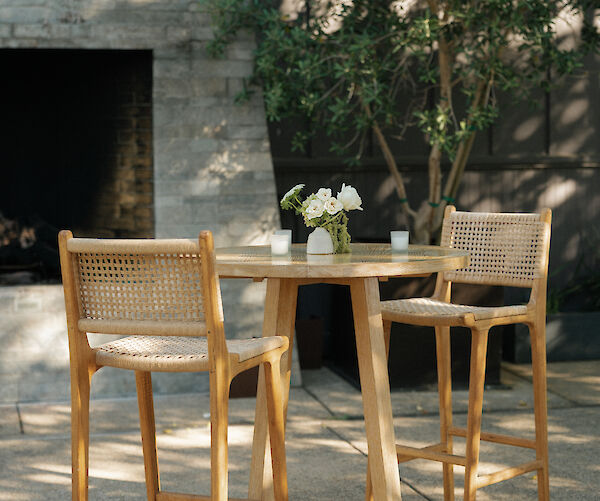 The image shows an outdoor scene with a wooden high table, two chairs, and a small vase with flowers, set against a garden background.