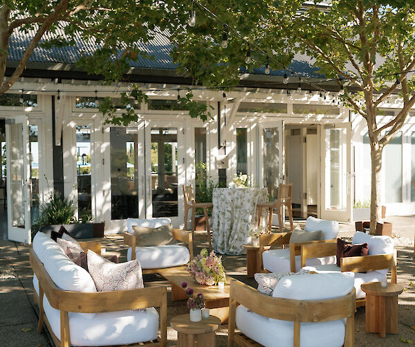 An outdoor seating area with wooden furniture and white cushions is set under trees, near a building with glass doors and windows.