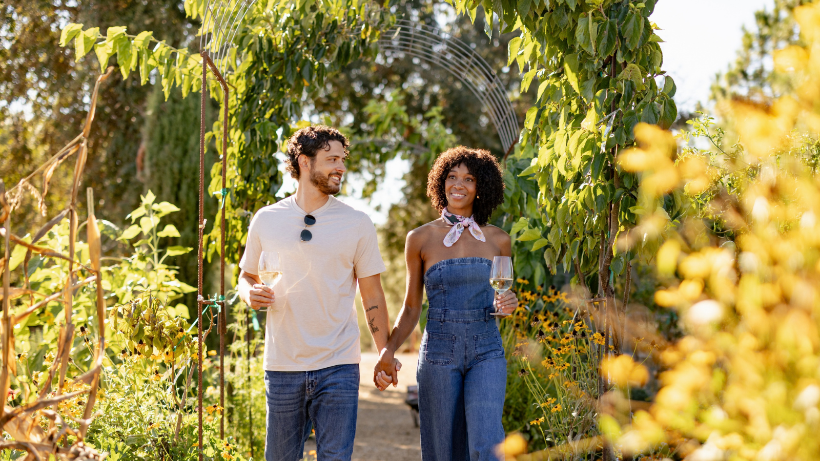 A couple is walking hand in hand through a lush garden path, each holding a drink, surrounded by vibrant greenery and flowers.