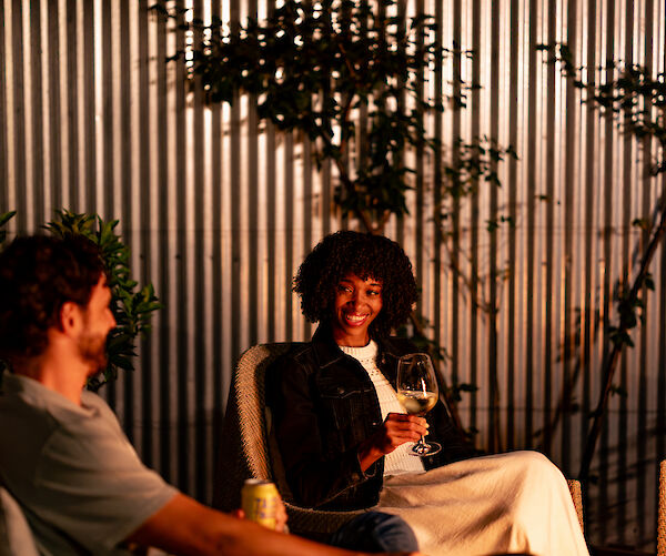 Two people are sitting outside at night, talking and holding drinks, with a plate of snacks on the table nearby.