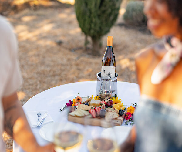 A couple toasts with white wine near a table displaying snacks and a bottle of wine in an outdoor setting.