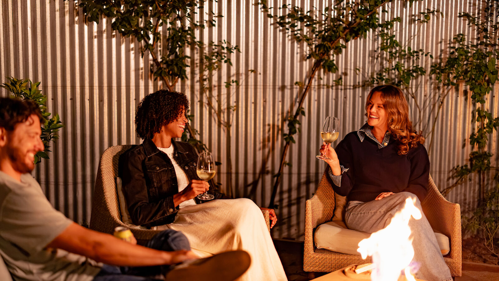 Three people sit around a fire pit outdoors, chatting and holding drinks, surrounded by greenery against a corrugated metal wall.