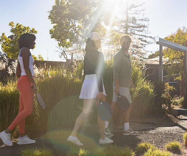 Three people in athletic wear walk outside with paddles in hand, likely heading to play a sport, under bright sunlight.