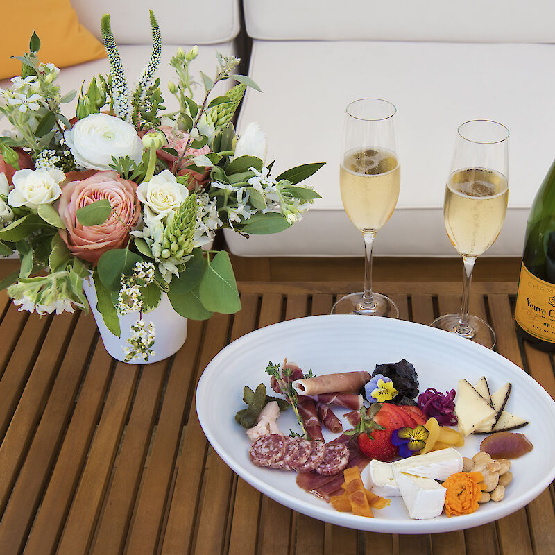 A table with a floral arrangement, a wine bottle, two filled wine glasses, and a charcuterie plate featuring meats, cheeses, and fruits.
