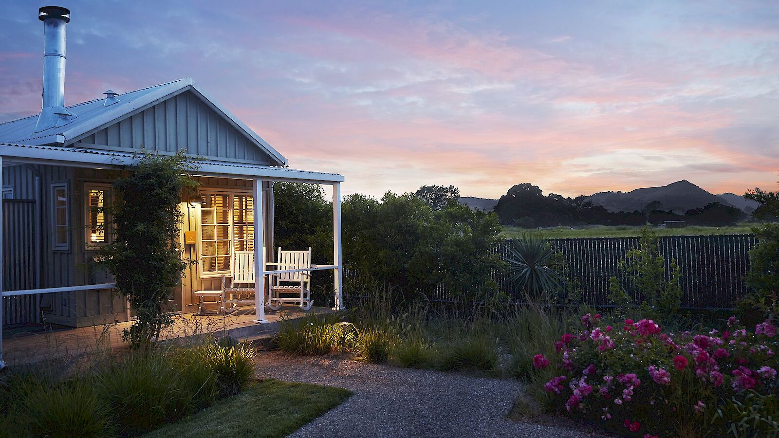 A cozy cottage with a lit porch sits in a garden, surrounded by greenery and flowerbeds, under a picturesque sunset sky with distant hills.