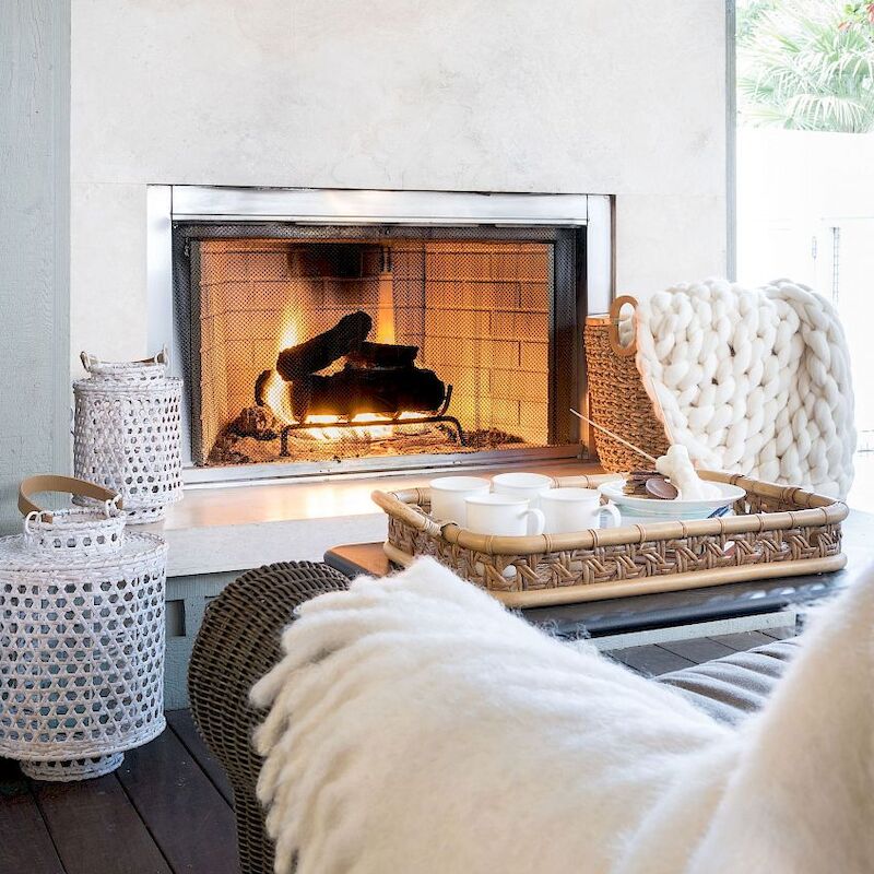 A cozy living room with a lit fireplace, wicker furniture, white and beige decor, blankets, and a tray holding cups and items on a table.
