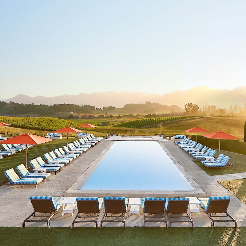 An outdoor swimming pool surrounded by lounge chairs and umbrellas, set in a scenic landscape with vineyards and mountains at sunset.
