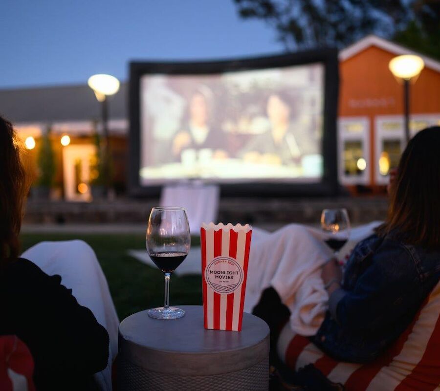 People watching an outdoor movie on a large screen, with wine glasses and popcorn in the foreground.