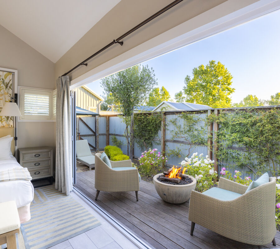 A cozy bedroom opens to a patio with chairs and a fire pit, surrounded by greenery and flowers, under a clear blue sky.