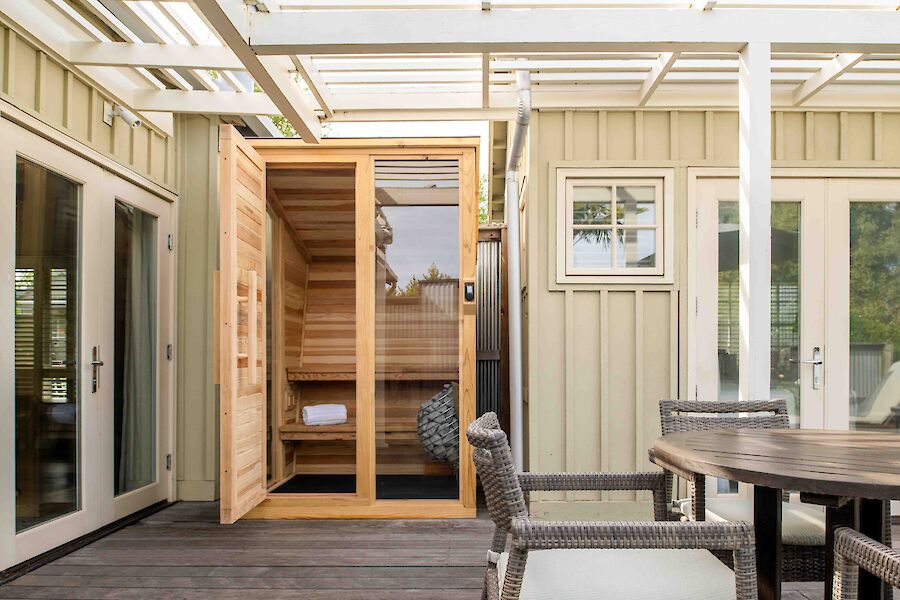 The image shows a patio with a wooden sauna, doors open, surrounded by a table, chairs, and a partially covered pergola.