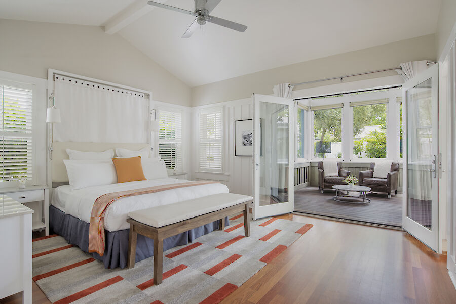 This image shows a modern bedroom with a bed, bench, and adjacent seating area through open double doors, with lots of natural light.