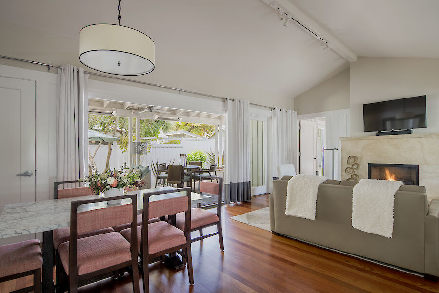 A cozy living-dining area with a fireplace, TV, and pink chairs. A table set with flowers and open patio doors complete the scene.