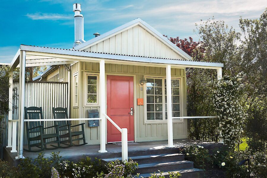 A small house with a pink door, a porch, two rocking chairs, and surrounded by greenery under a blue sky.