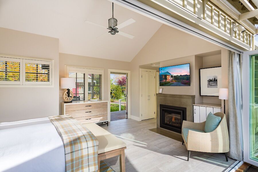 A bright, modern bedroom with a bed, fireplace, chair, and large windows opening to a balcony. There's art on the walls and a ceiling fan.