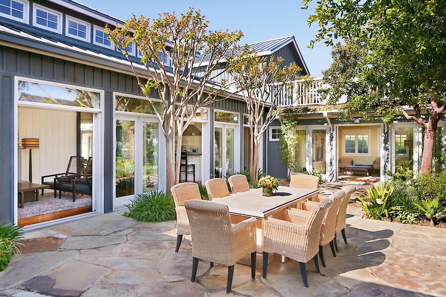 A patio with a table, wicker chairs, and surrounding greenery outside a house with large windows.