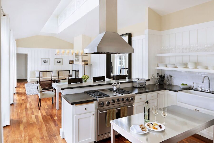 The image shows a modern kitchen with a stainless steel island, stove, white cabinetry, and a dining area. Light streams in from large windows.