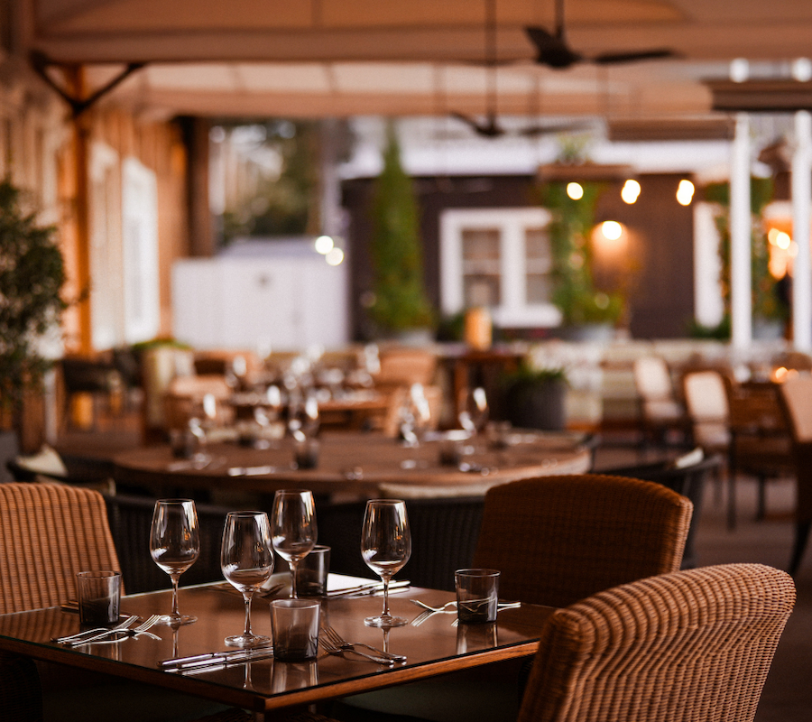 The image shows a cozy outdoor restaurant setting with neatly arranged tables, chairs, and ambient lighting.