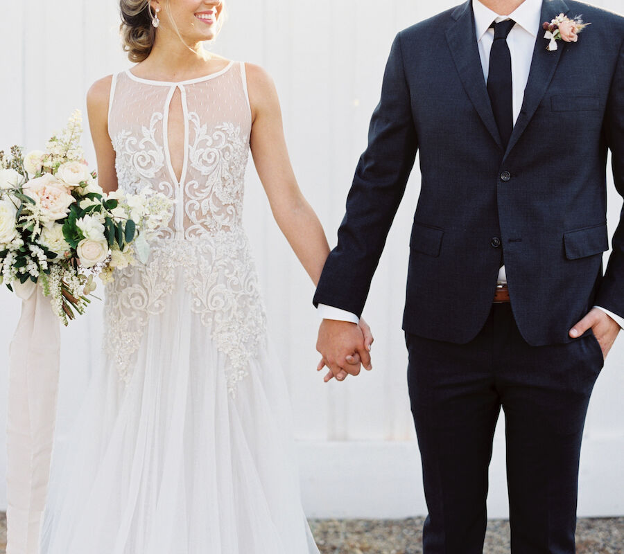 A couple is holding hands; one in a wedding dress with a bouquet, the other in a dark suit.