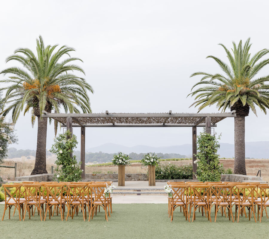 An outdoor wedding setup with wooden chairs facing a pergola adorned with flowers, framed by palm trees on a grassy area.