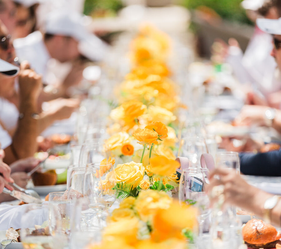 A long table set for a meal, decorated with bright yellow flowers, with people dining on both sides in an outdoor setting.