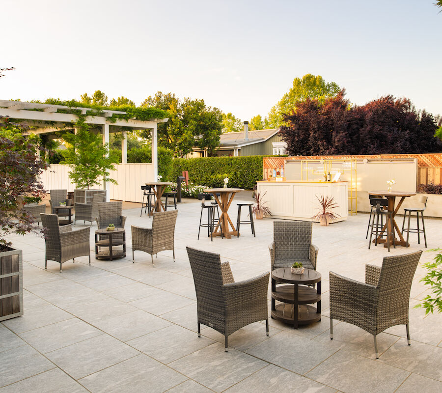 The image shows a spacious outdoor patio with wicker furniture, tables, and a bar area, surrounded by greenery and trees in the background.