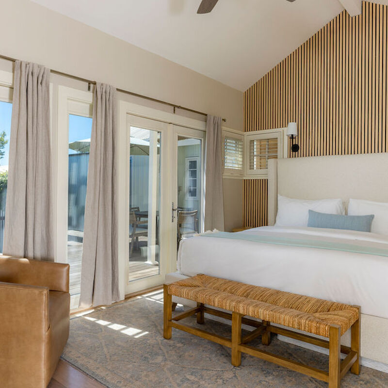 A bedroom with a wooden slat wall, large bed, leather chair, and bench, filled with natural light from glass doors.