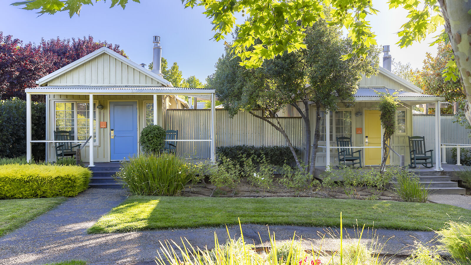 The image shows two quaint cottages with porches, surrounded by lush greenery and garden landscaping under clear skies.