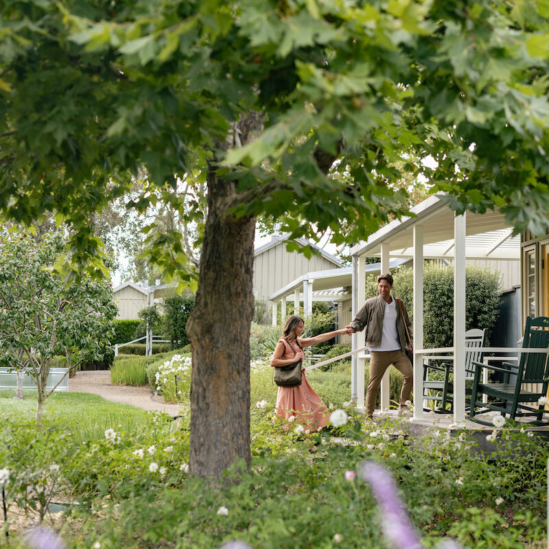 A couple walking near a quaint porch surrounded by trees and flowers, creating a peaceful and scenic atmosphere.
