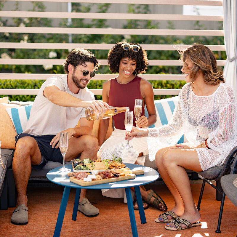 Three people sit outdoors on a patio, sharing food and drinks, appearing joyful and relaxed, with a modern seating area around them.