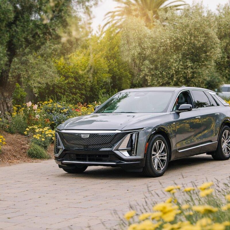 A sleek black car is parked on a sunny, flower-lined cobblestone path surrounded by lush greenery and trees.