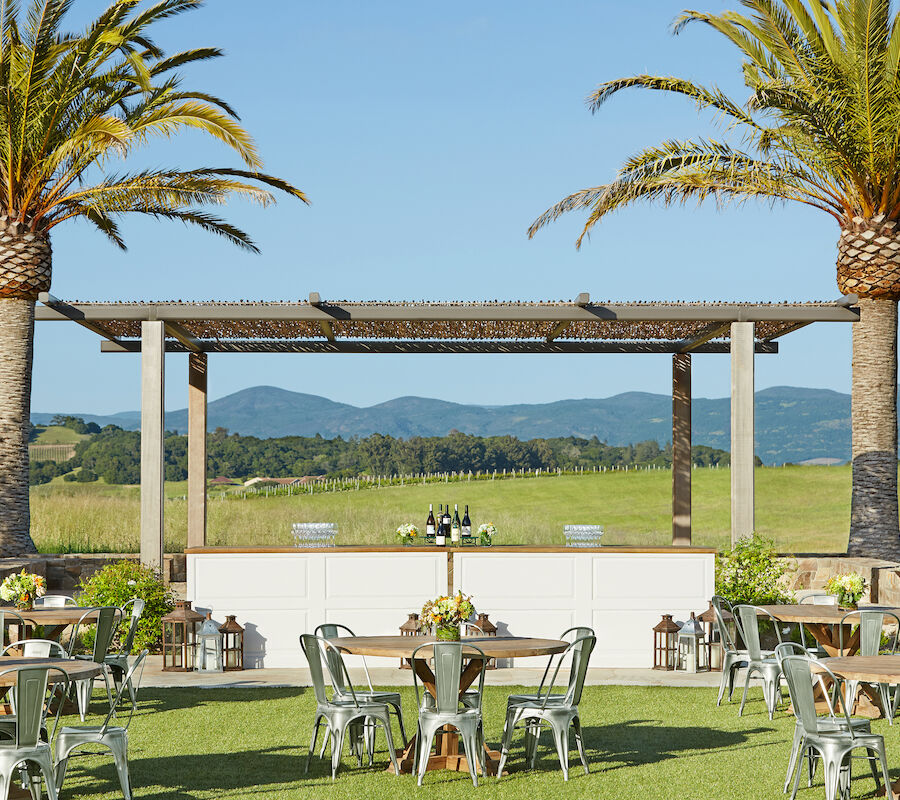 An outdoor event setup features tables and chairs under palm trees with a bar in the background, all against a scenic backdrop of rolling hills.