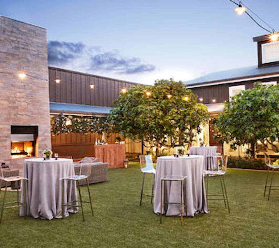 An outdoor area with tables draped in linens, chairs, a fireplace, and trees, set for an event, with string lights creating a cozy ambiance.