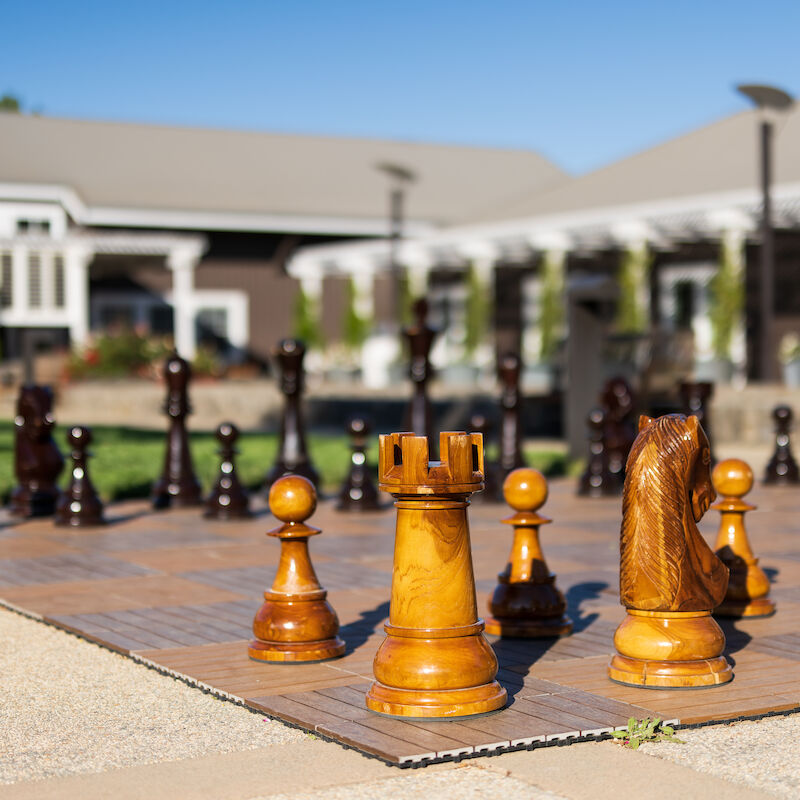A large outdoor chessboard with oversized wooden chess pieces placed on it, set in a garden with a building in the background.