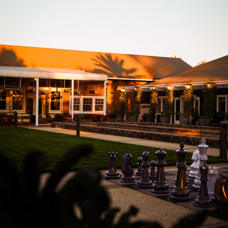 The image shows a large outdoor chess set in front of a building with a warm sunset glow, surrounded by greenery.