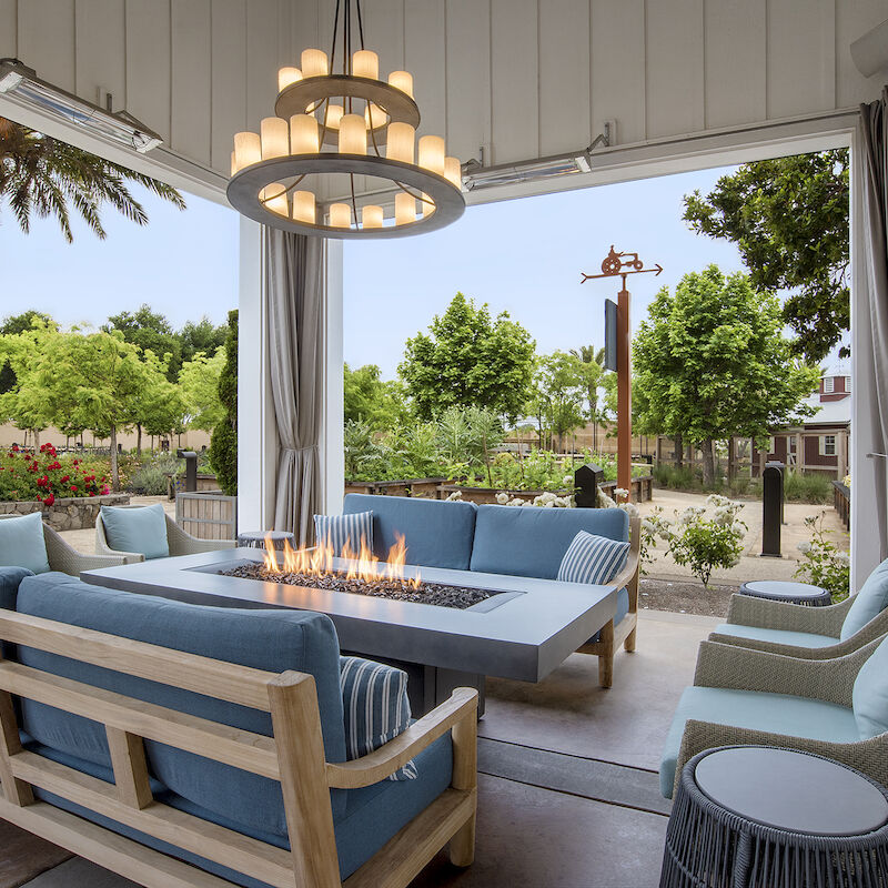 An outdoor seating area with a fire pit, surrounded by blue cushioned furniture and a chandelier, overlooks a garden and palm trees.