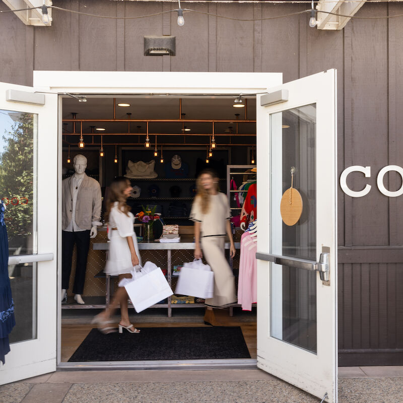 Two people walking out of a clothing store with shopping bags. Garments are displayed on racks, and mannequins are visible inside.