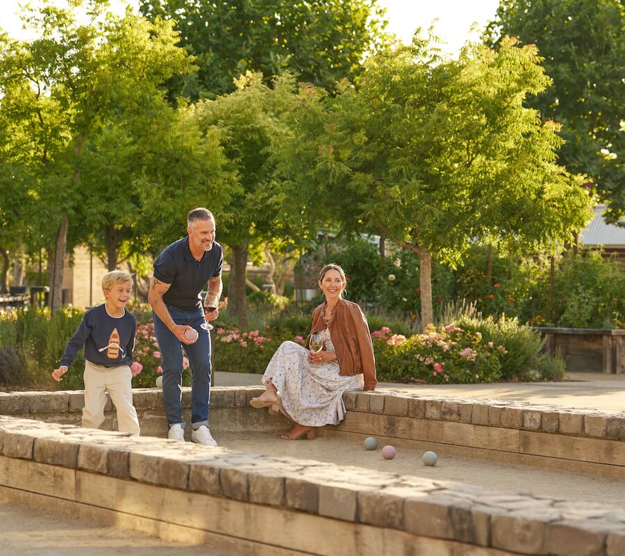 A family is playing bocce ball in a sunny outdoor park with trees and flowers surrounding them.