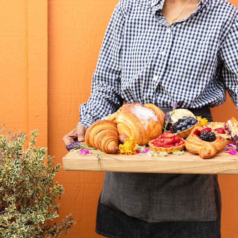 A person holds a wooden tray full of pastries, including croissants and fruit-topped delights, against an orange background.