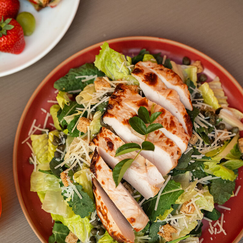 A salad with sliced grilled chicken, lettuce, cheese, and herbs is on a red plate. A side plate with strawberries is partially visible.