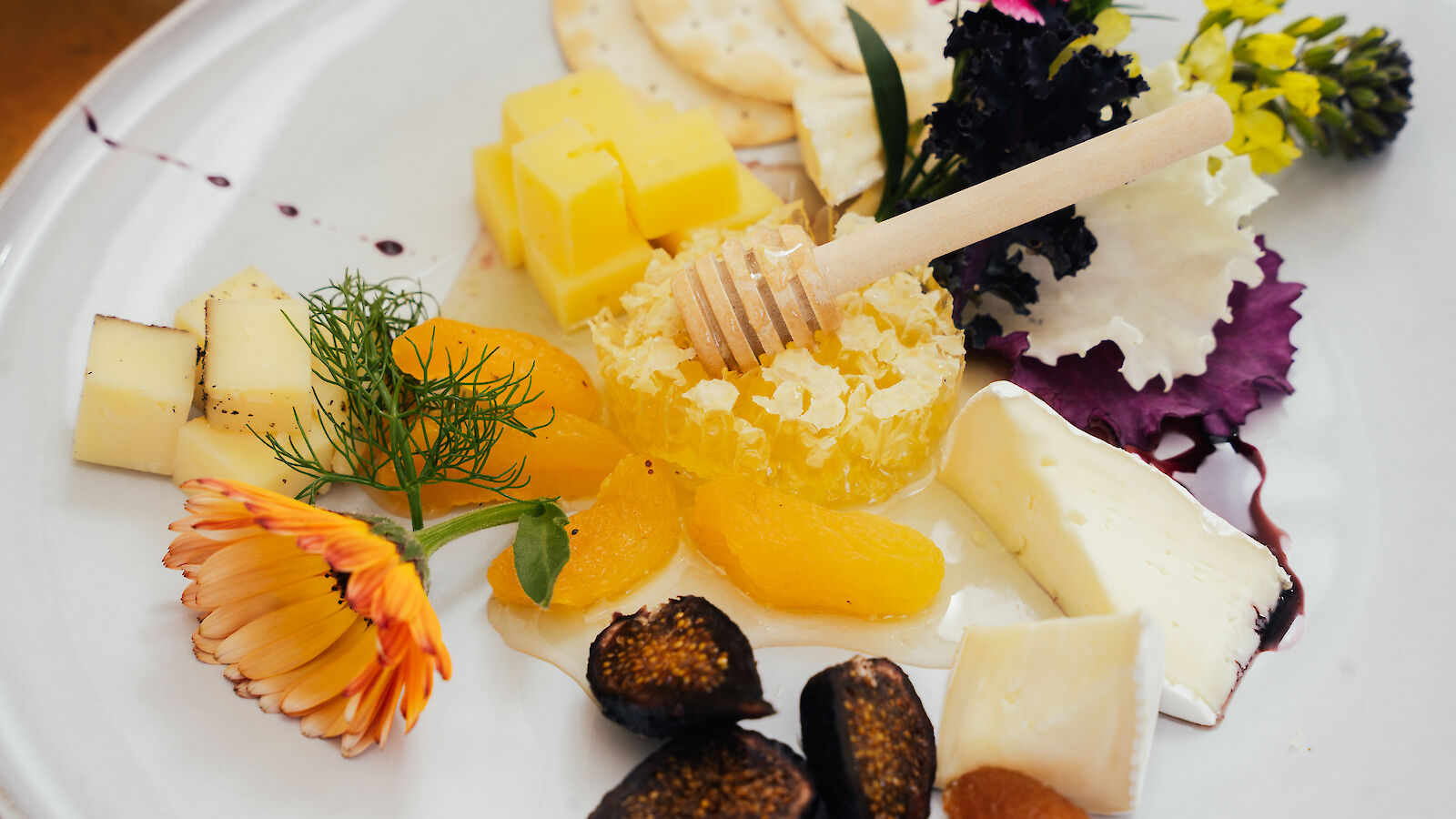 A cheese platter with various cheeses, sliced fruits, crackers, honeycomb with a dipper, and edible flowers is artfully arranged on a plate.