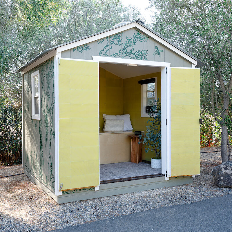 A small shed with open yellow doors, revealing a cozy interior with cushions and a window, surrounded by trees and a gravel path.