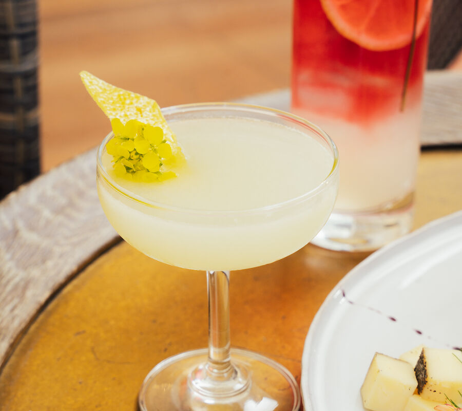 Two cocktails on a table, one with citrus garnish and the other with fruit slices; part of a plated dish is visible.