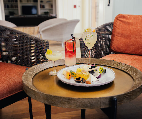 A round table with two orange chairs holds three cocktails and a plate of assorted cheeses and fruits, creating a cozy lounge setting.