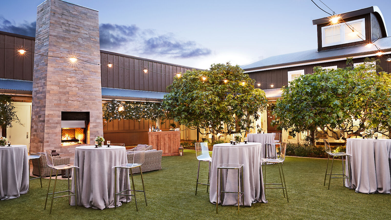 An outdoor venue with cocktail tables, string lights, trees, and a large brick fireplace, set against a modern building.