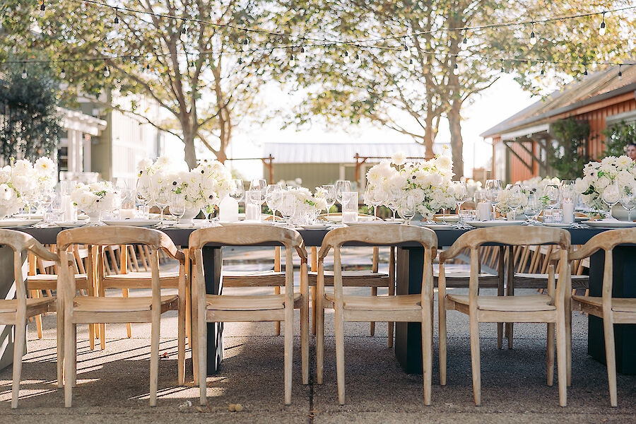 An outdoor dining setup with wooden chairs and tables adorned with white floral arrangements under trees.