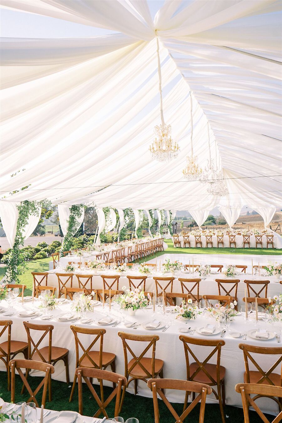 An elegantly decorated outdoor event setup under a white canopy with rows of wooden chairs and tables adorned with flowers and tableware.