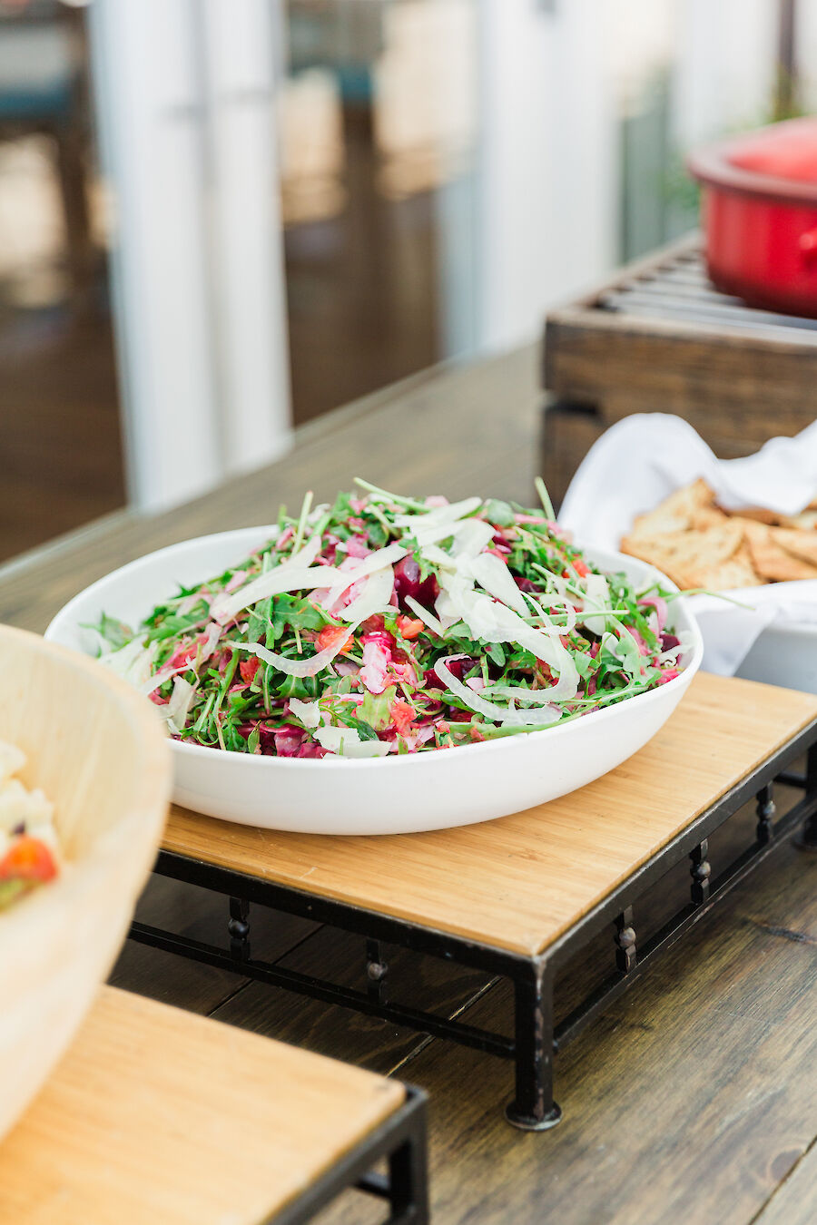 The image shows a bowl of fresh salad with greens and radishes on a wooden stand, surrounded by other dishes on a rustic table.