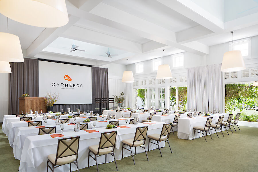 A modern conference room with tables, chairs, a projector screen, and natural light through large windows.