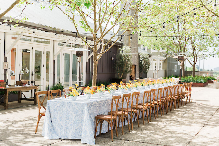 An outdoor dining setup with a long table, floral centerpieces, wooden chairs, and string lights under trees beside a building.